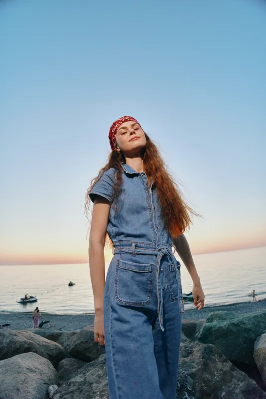 lifestyle woman in modern boho-western denim outfit stands confidently on rocky shore at sunset, film color capturing natural mood and street style vibe