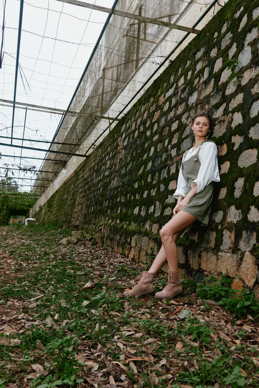 Fashionable young model in a green outfit posing against a rustic stone wall, captured in a natural setting with vibrant greenery, exuding a confident and serene emotion