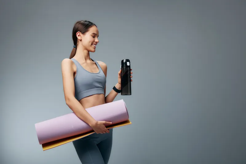 Fit woman in sportswear stands with a yoga mat under her arm and a water bottle in hand, posing in a minimalist studio for a fitness and sport lifestyle concept.