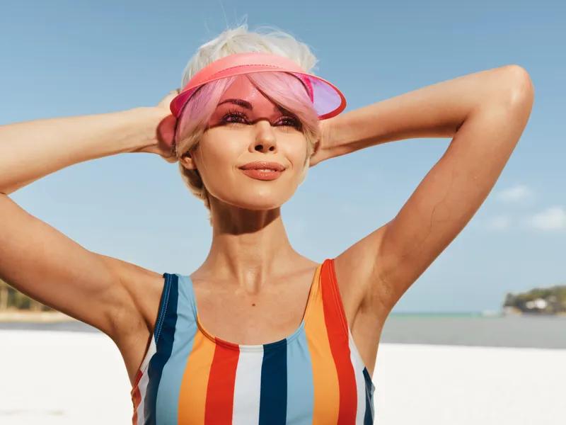 Portrait of a confident woman on a sunny beach wearing a striped swimsuit and pink visor, posing with hands behind head against a bright sky and blue sea.