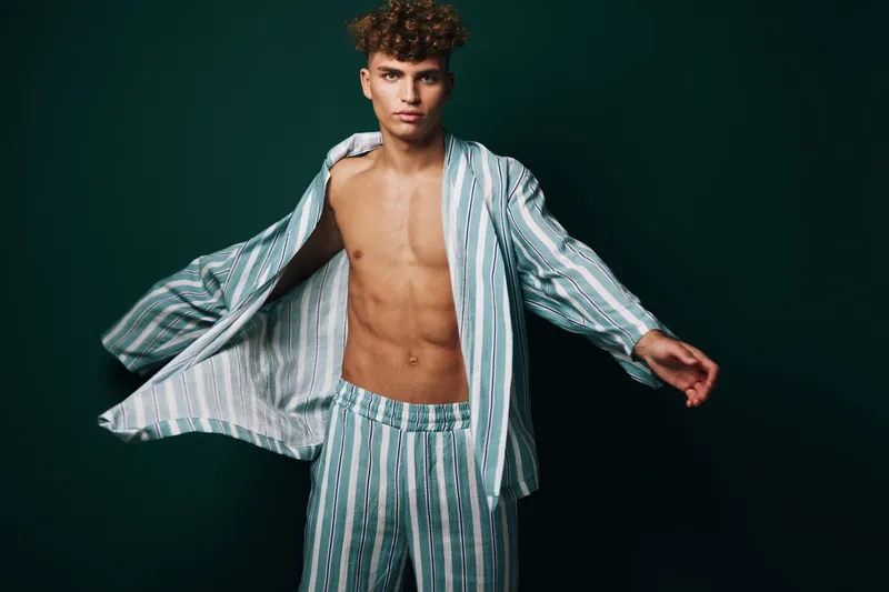 Young man with curly hair wearing striped pajamas posing with open shirt and confident expression on dark background for fashion and lifestyle concept.