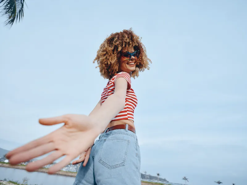 Joyful young woman extends an open hand toward the camera on a sunny day, wearing striped top and denim shorts, capturing carefree spirit and playful energy.