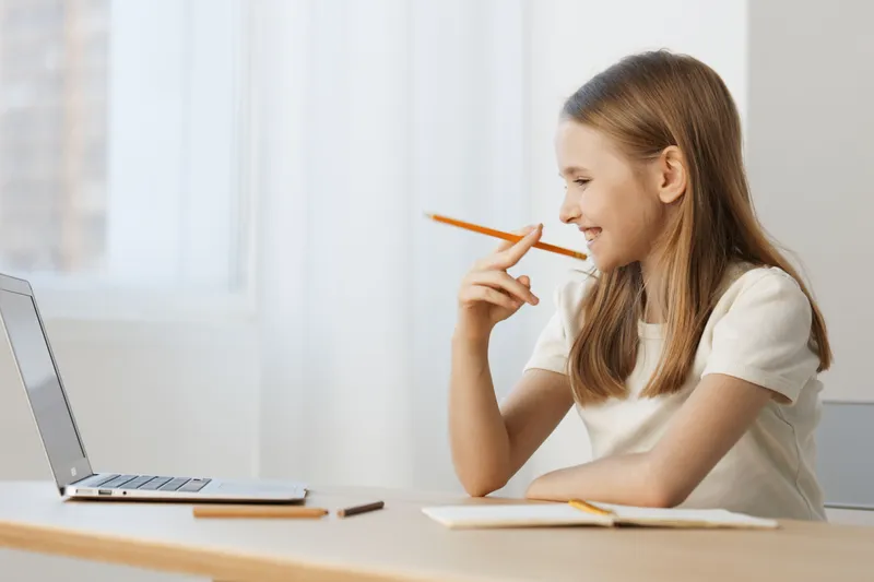 Thoughtful girl smiling while studying online, engaged with a laptop in a bright, modern room, showcasing determination and focus