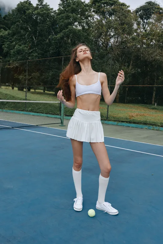 Stylish young female athlete posing confidently on a tennis court, wearing a sports crop top and pleated skirt with a vibrant green backdrop