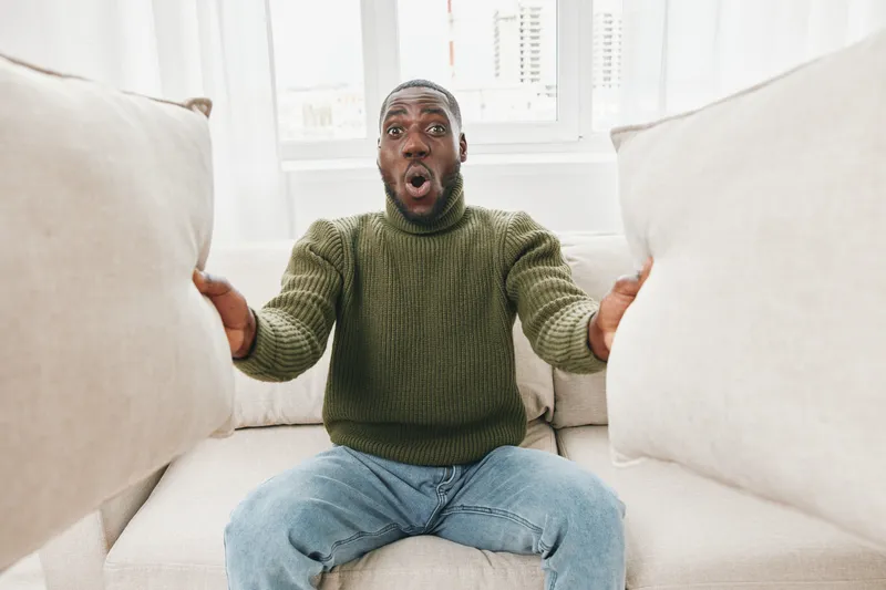 Surprised young Black man sitting on a couch holding large cushions with a shocked expression, wearing a green sweater and jeans in a bright living room