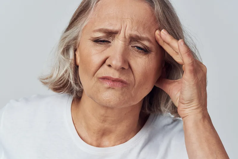Пожилая женщина держится за голову, страдая от мигрени и головной боли, в поисках лечения