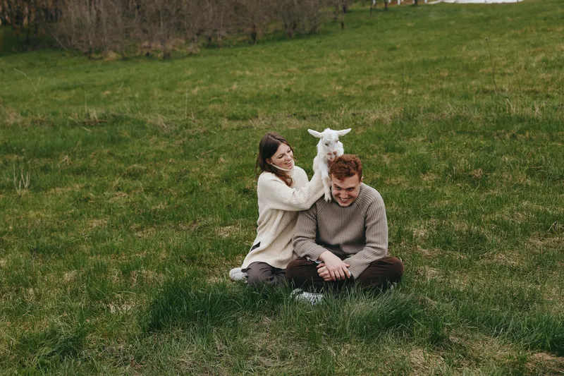 Happy couple sits on a grassy field with a playful goat kid between them, sharing a gentle moment in cozy sweaters, natural light, candid lifestyle portrait outdoors, loving interaction and rural