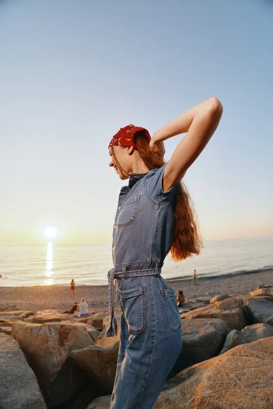 Lifestyle woman in modern boho-western denim street style stretching at sunset on rocky beach with film color warm tones and relaxed mood.