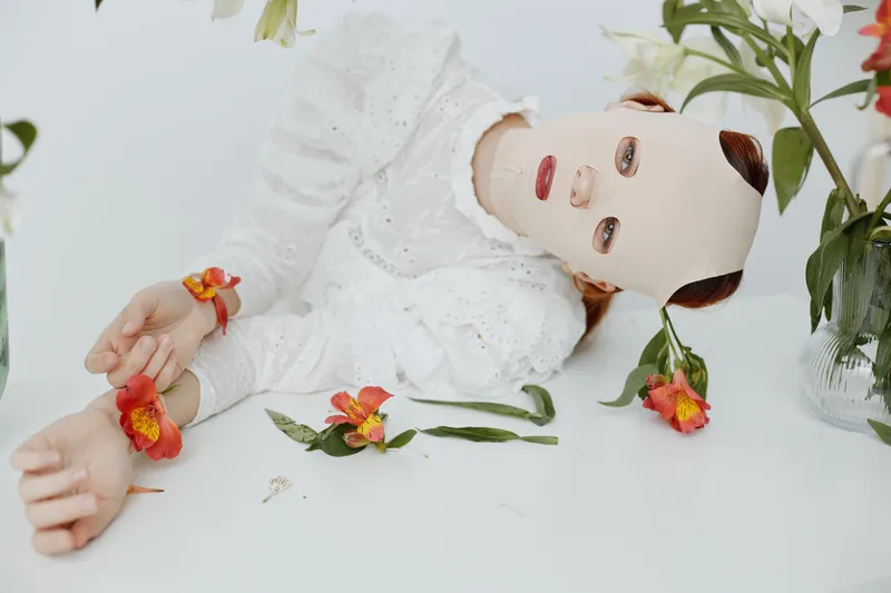 A surreal concept photo featuring a model with a mask, lying among flowers, evoking emotions of mystery and beauty in a soft, pastel color palette