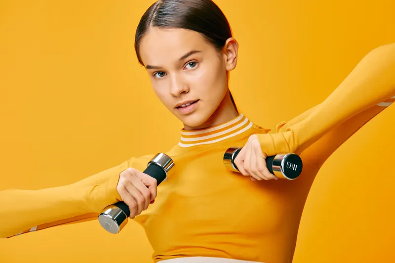 A fitness woman lifts dumbbells in each hand, performing a focused workout in a vibrant orange studio. This athletic training session emphasizes strength, balance, and determination.