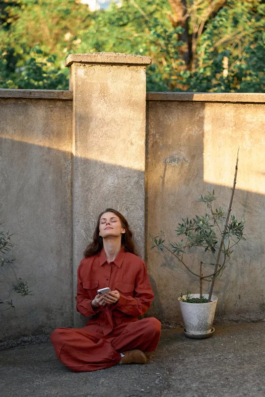 Woman in red outfit sits cross legged beside a sunlit wall, eyes up, holding a phone. Calm outdoor mood, casual lifestyle, natural light, intimate moment, simple everyday scene