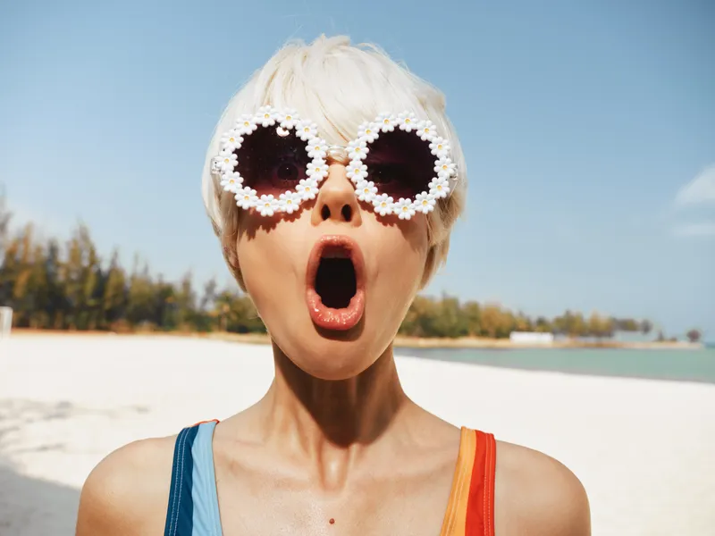 Surprised woman on a sunny beach wears flower sunglasses, open mouth expression, bold fashion moment, capturing playful summer style and dynamic personality.