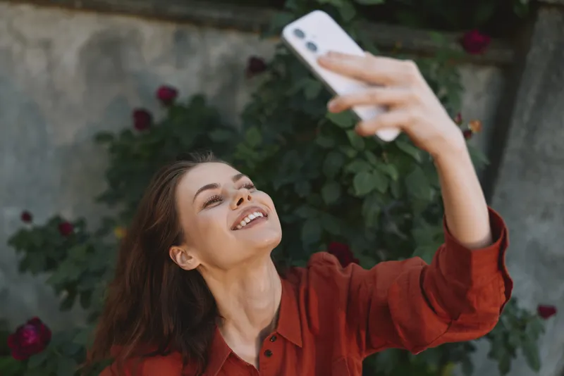 Woman lifestyle selfie outdoors smiling while holding smartphone, captures joyful moment in red shirt, casual pose against floral backdrop, bright natural light, candid portrait for social media