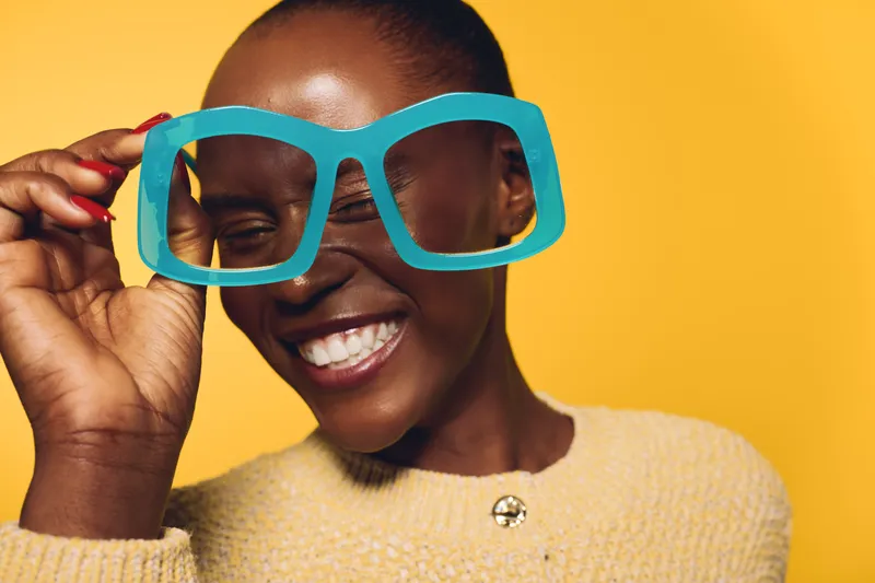 Happy black woman with dark skin smiles joyfully holding oversized blue glasses on yellow background. Concept of positive emotions, fashion accessory, and playful mood for African ethnicity.
