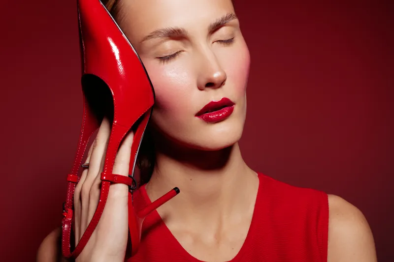 Close-up of a woman with closed eyes holding a red high heel shoe near her face. Red background and red dress create a bold fashion and beauty concept.