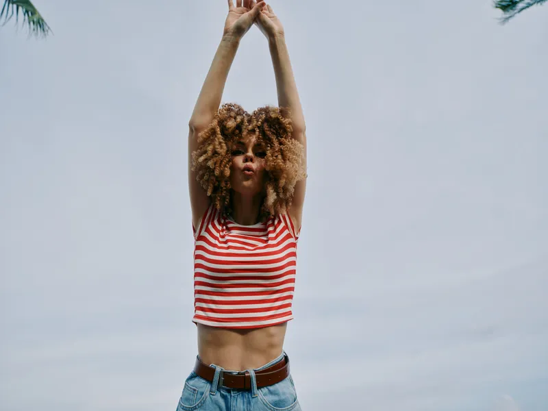 Young woman stands outdoors with arms raised above her head, wearing a red and white striped crop top and denim jeans, set against a bright blue sky.