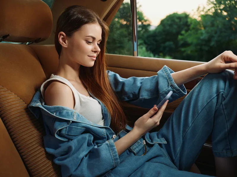 Young woman in denim outfit enjoying her smartphone in a car, showcasing a relaxed mood with a scenic green background.