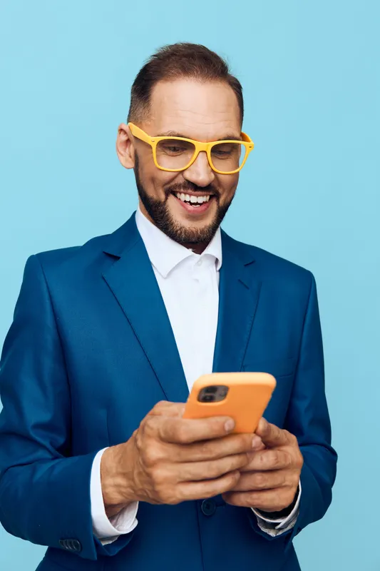 A professional man in a sharp suit stands against a solid colored background, exuding confidence and focus while glancing at a mobile device, ready to engage in business tasks.