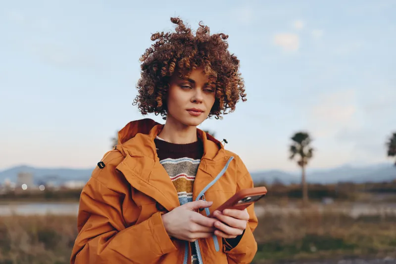 Woman lifestyle outdoors, curly hair and orange jacket, holding a smartphone in hand, calm expression, soft daylight over a rural field, distant hills and sky, candid moment