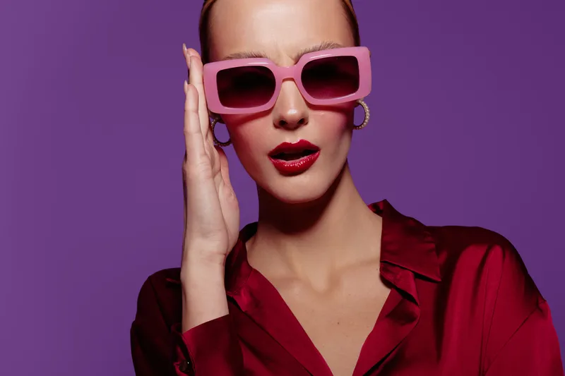 Stylish woman in pink sunglasses and red shirt poses against purple background. Fashion portrait with bold makeup and confident expression. Concept of glamour, style, and modern beauty.