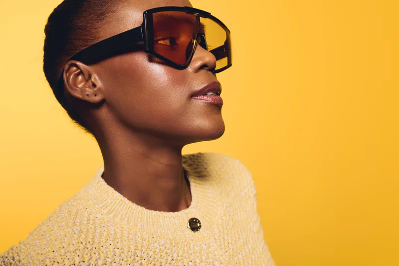 Side profile of confident black woman with dark skin wearing large sunglasses and yellow sweater against bright yellow background. Concept of fashion, style, and modern beauty.