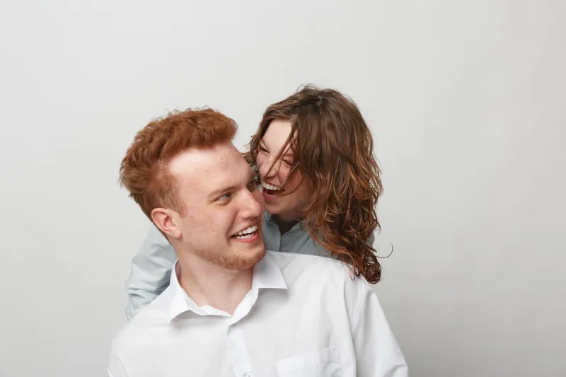 Happy young couple with red hair and casual white shirts laughing together on light background. Concept of friendship, joy, and positive emotions in relationships and lifestyle.