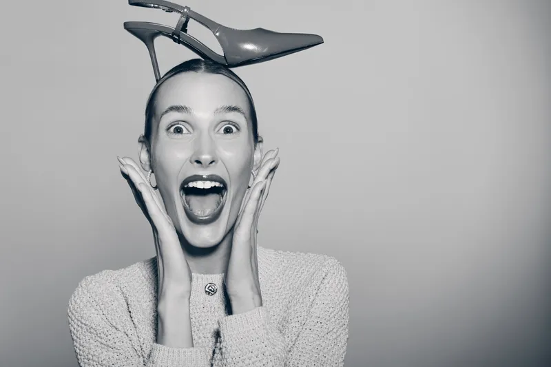 White woman with fair skin and red lipstick looks surprised with open mouth and holds high heel shoe on head in studio. Concept of fashion, fun, and emotion.