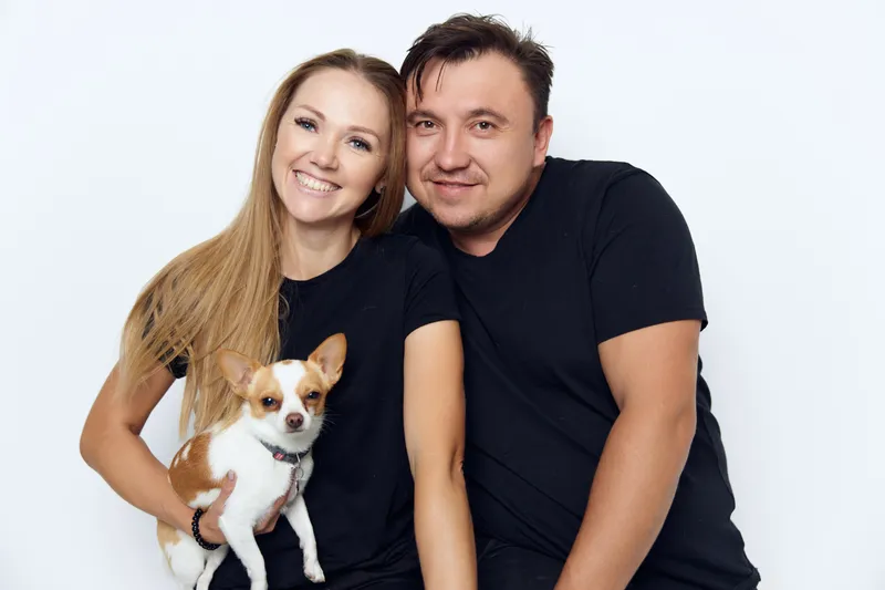 Счастливая супружеская пара обнимается в черных футболках в фотостудии, держа в руках маленькую собаку