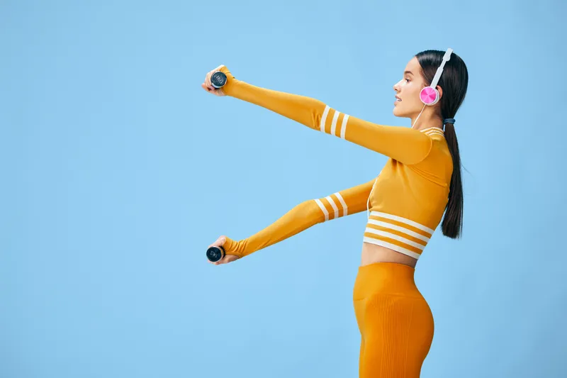 Woman fitness, fitness routine and dumbbell workout with arm extension in a vibrant studio. Athletic female athlete wearing orange outfit and headphones performing a dynamic arm exercise against a