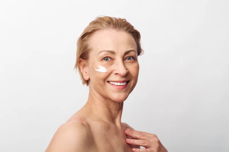 Smiling mature woman with short blonde hair applies white face cream on cheek, showing healthy skin and natural beauty. Skincare, wellness, and self-care concept with soft lighting on white