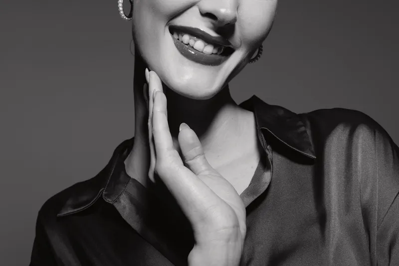 Smiling Caucasian woman with light skin touches face with hand, wearing dark satin shirt in studio. Concept of beauty, skincare, and happiness.