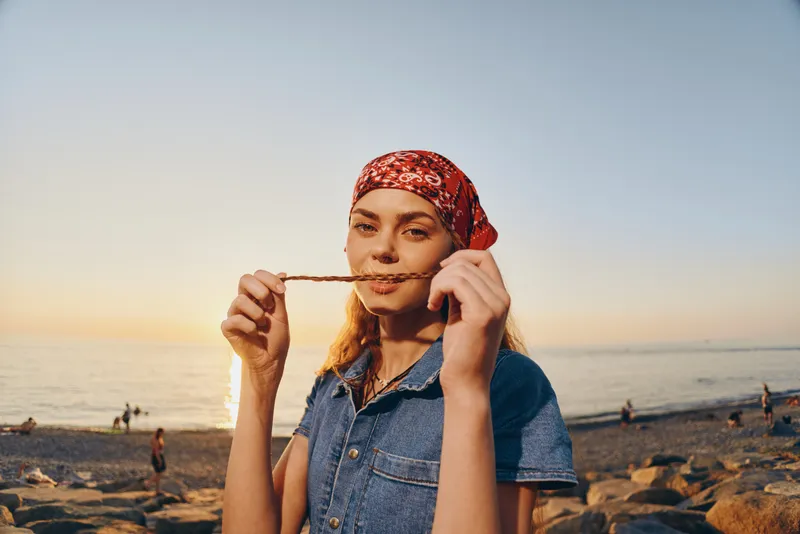 smiling woman wearing red headscarf and denim shirt playing with braided hair at sunset on rocky beach with calm sea and clear sky in background