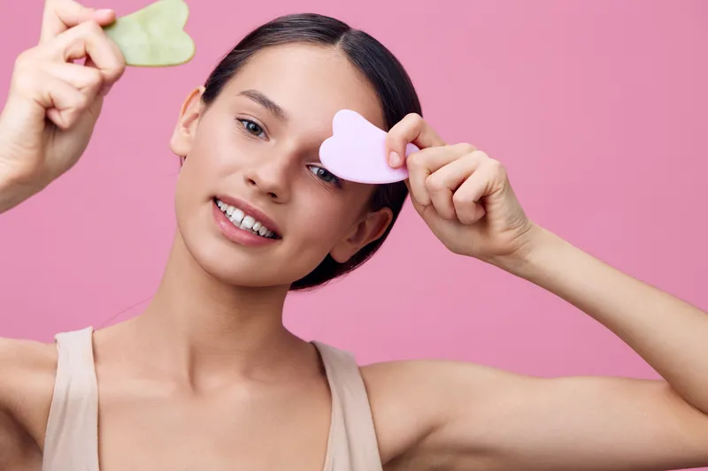 Young woman holding heart shaped beauty tools, showcasing a joyful expression against a soft pink background, representing skincare and self care concepts