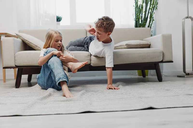 Playful children indoors, a smiling boy and girl engaging in a friendly game on a carpet in a bright living room with modern furniture and natural light.