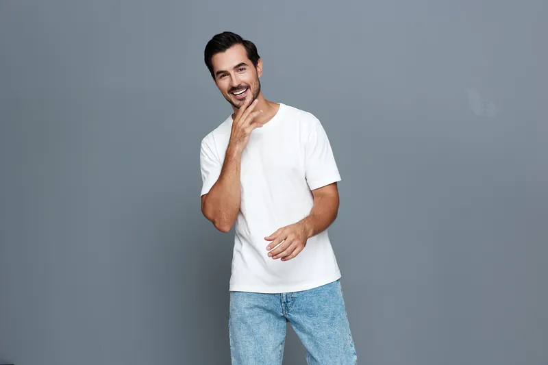 Smiling man wearing casual white t-shirt and jeans, looking at the camera with a joyful expression, standing against a plain gray background. Lifestyle concept.