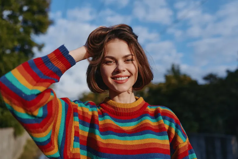 Young woman, smiling woman in a rainbow sweater outdoor portrait with short hair and casual outfit, mid twenties female model smiling, hand in hair, bright lifestyle fashion.