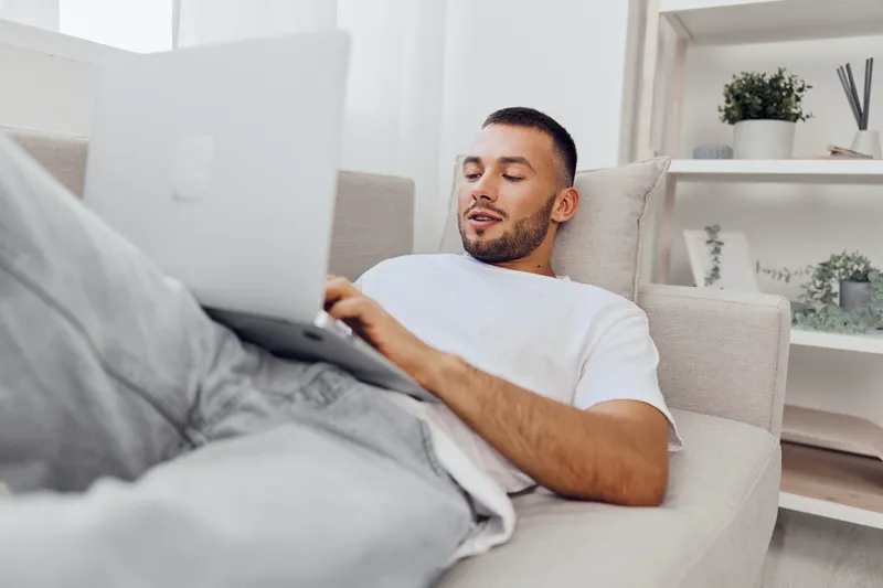 Relaxed man using a laptop on a sofa, showcasing a modern home environment filled with natural light. Perfect for wellness and remote work themes.