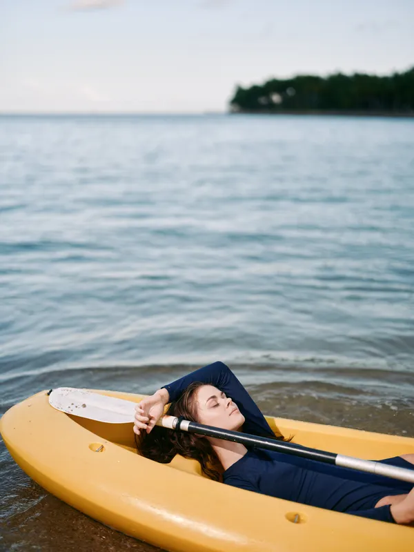 Relaxed woman lying in a yellow kayak on calm water with a peaceful sky and distant shore, enjoying a tranquil moment during a warm day outdoors.