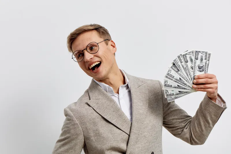 happy young man holding dollar bills in hand smiling wearing glasses beige jacket casual outfit isolated on white background financial success concept