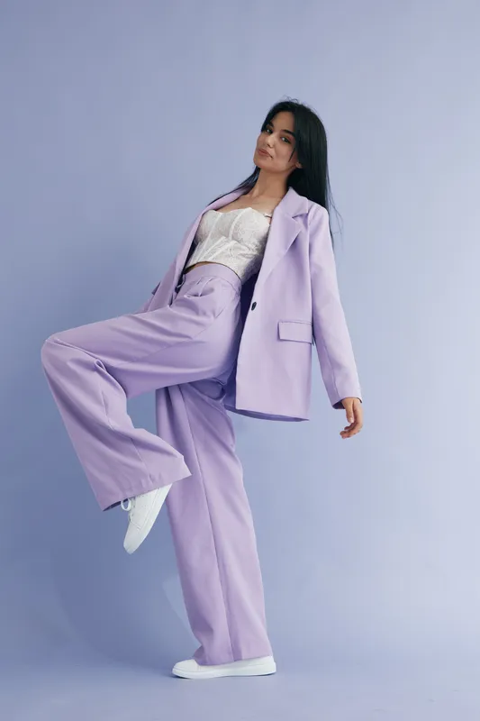 woman in lilac suit standing on one leg posing confidently in studio with soft purple background and casual white shoes, styled in modern fashion outfit