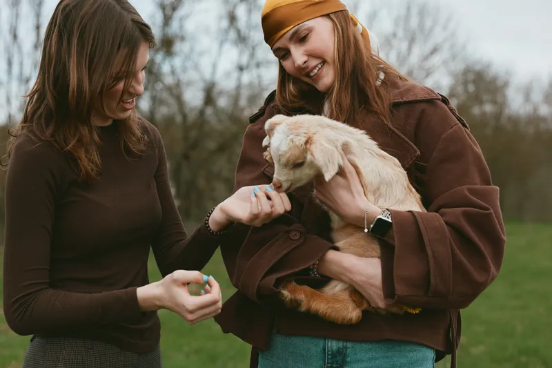 Two women outdoors admire a small goat kid, one offering a treat while the other gently holds the animal. Casual autumn outfits create a warm lifestyle moment focused on animals and friendship.
