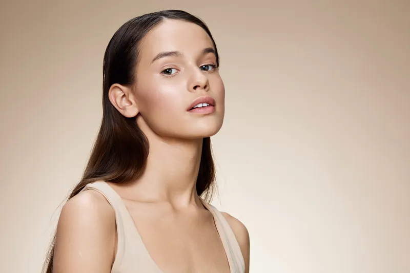 Young woman with long dark hair, natural beauty, soft makeup, neutral background, showcasing healthy skin and a serene expression, ideal for beauty themes