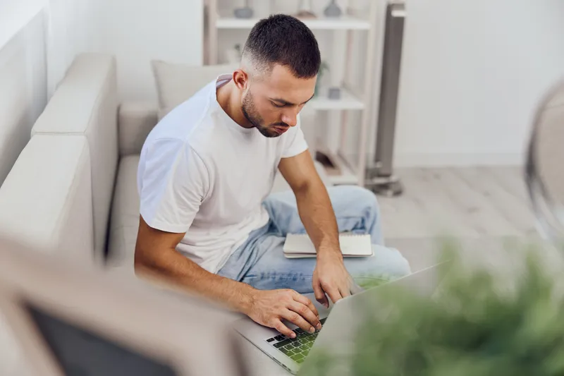 Focused man working from home on a laptop, wearing casual attire and surrounded by plants, creating a cozy workspace atmosphere. Concept: remote productivity.