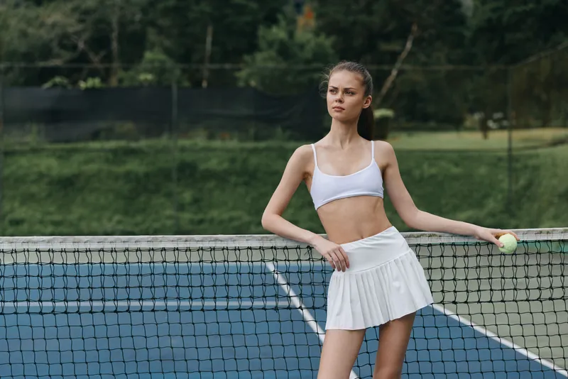 Young female tennis player in a white outfit poses confidently on a tennis court, showcasing athleticism and determination, surrounded by nature