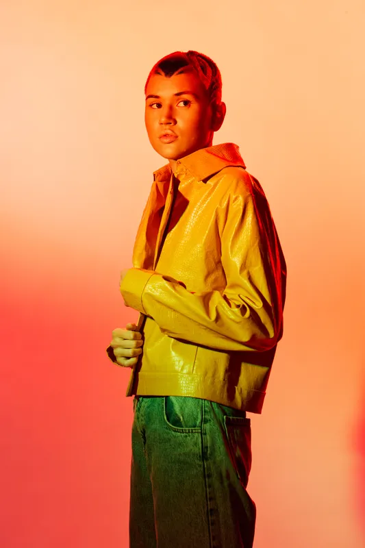 young woman in yellow leather jacket and blue jeans posing with serious expression against orange background under dramatic studio lighting