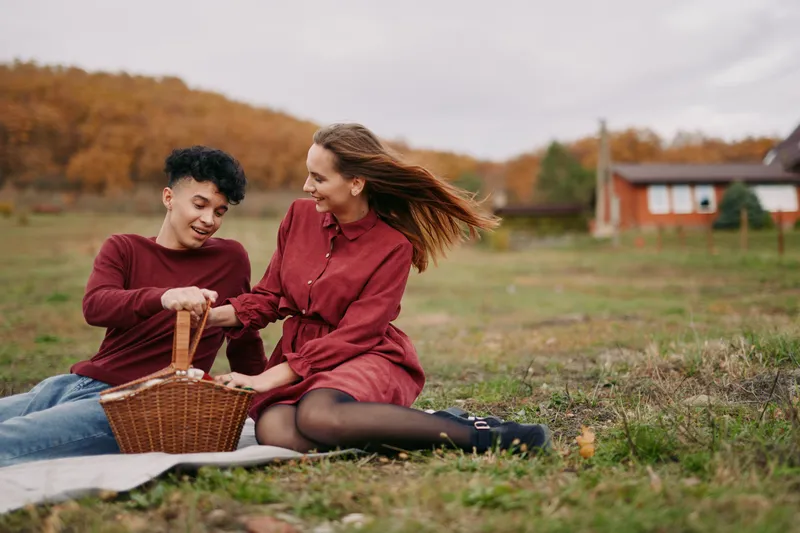 A candid outdoor picnic portrait of a young couple sharing a wicker basket on a blanket in autumn fields, natural smiles and gestures emphasizing authenticity and warm everyday connection.