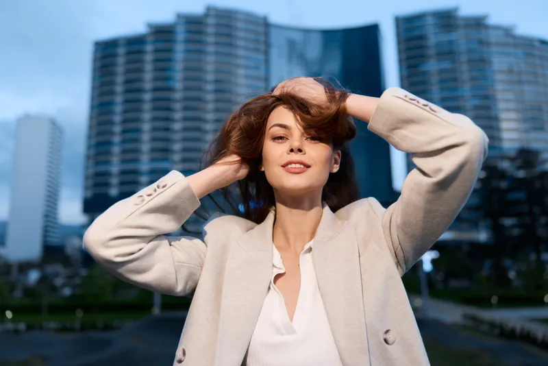 Confident woman in beige coat posing confidently in urban business district with modern skyscrapers and natural light