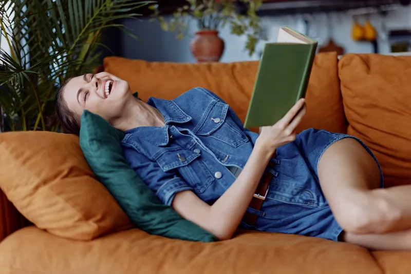 Женщина отдыхает дома, читая книгу на диване, перед которым стоит растение