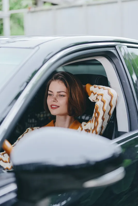 A portrait of a young woman driving a car, seated in the driver seat with a relaxed smile and confident gaze.
