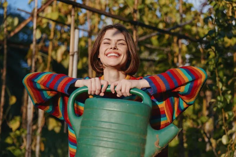 Woman with watering can in garden, smile and colorful sweater outdoors among green plants, happy gardener holding large green can, sunlight and joyful pose.